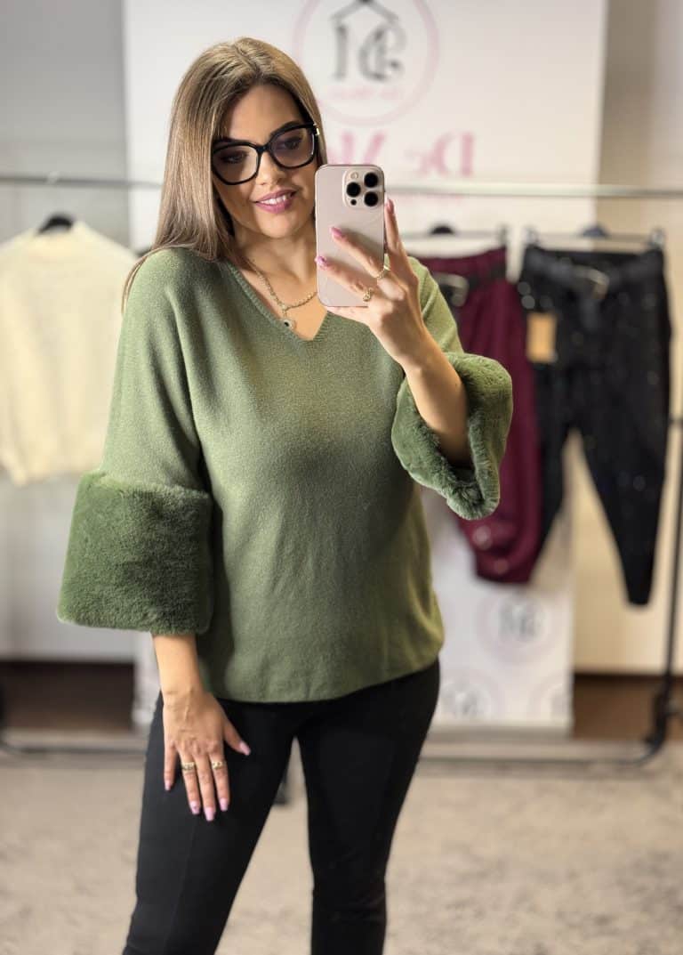 Női modell szőrmés ujjal kötött pulóvert visel, fekete nadrággal, fényképez selfie-t.