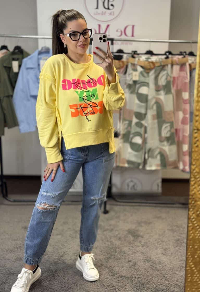 Női modell sárga oversized pulóvert visel, kék farmerrel és fehér sportcipővel.