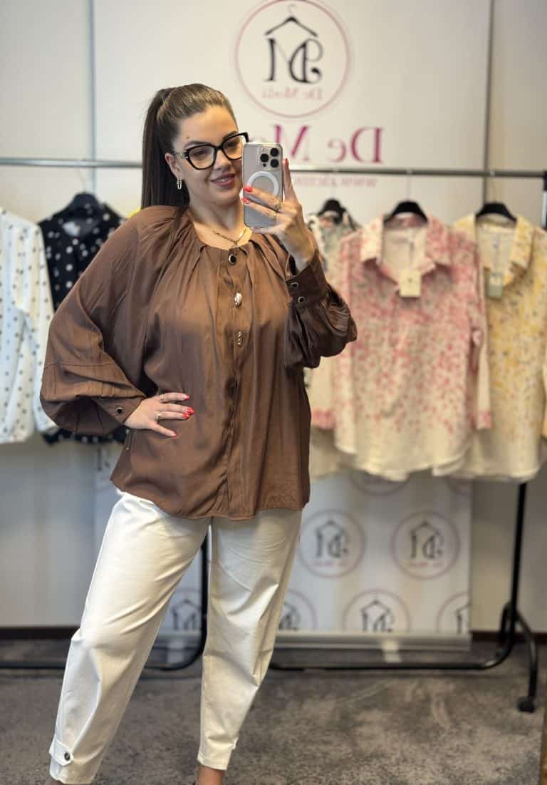 Női modell egy barna ingben és fehér nadrágban, háttérben ruhák a boltban.