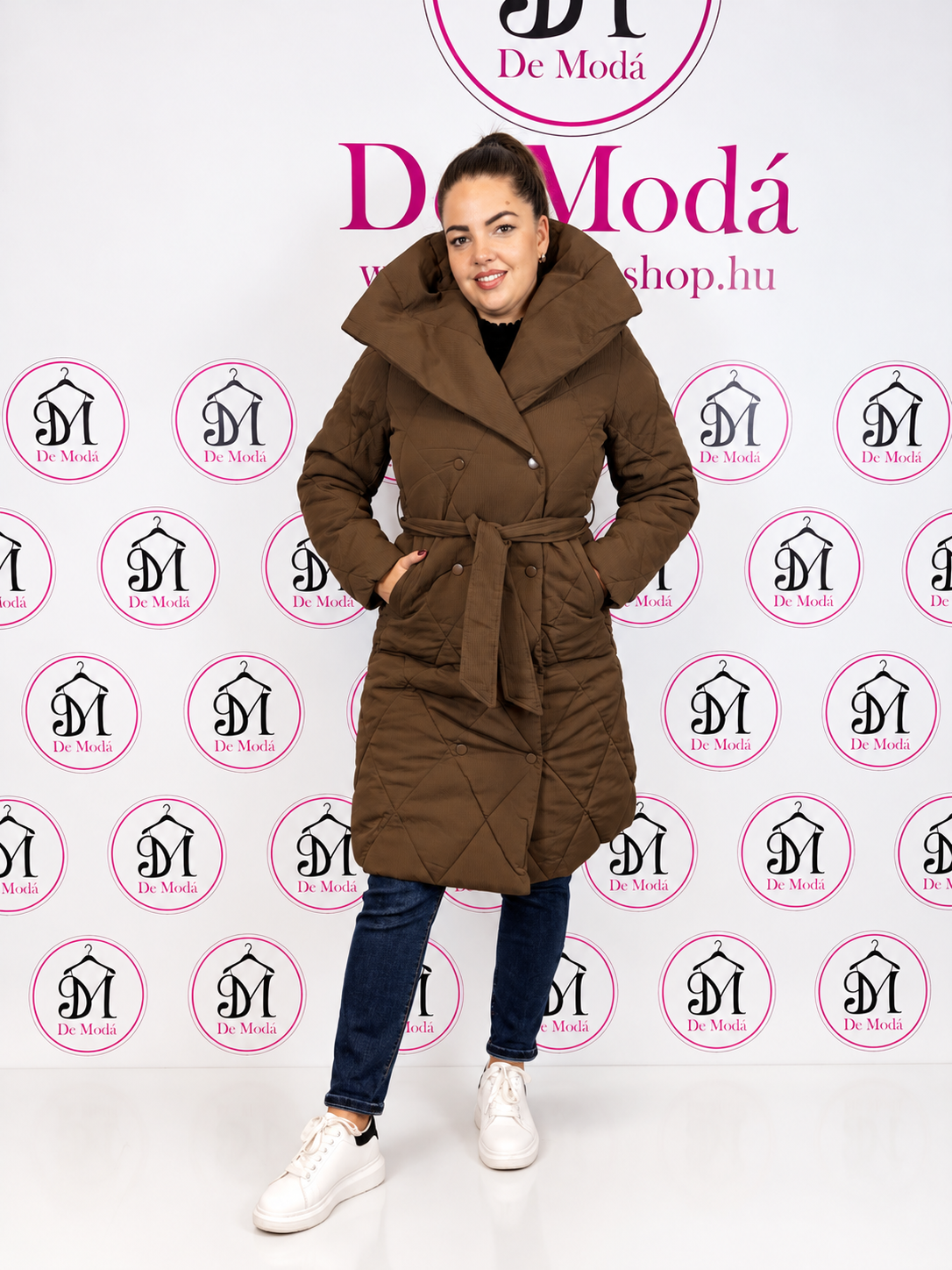 Nő modell egy barna steppelt kabátban, háttérben a De Moda logóval.
