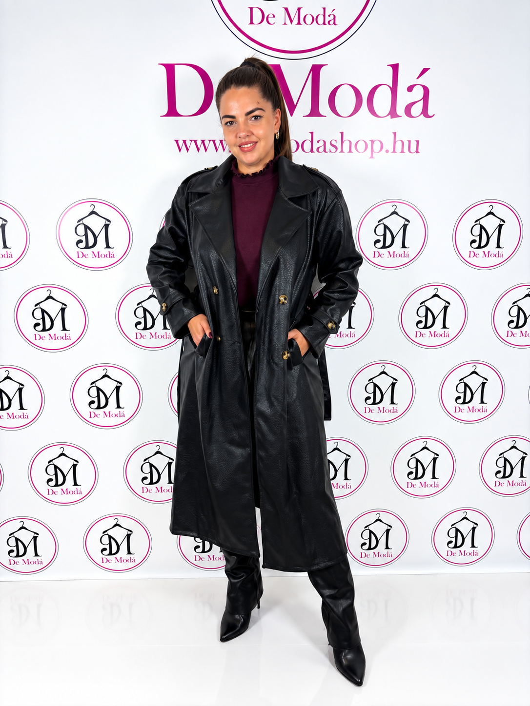Nő modell hosszú fekete műbőr kabátban, háttérben De Moda logóval.
