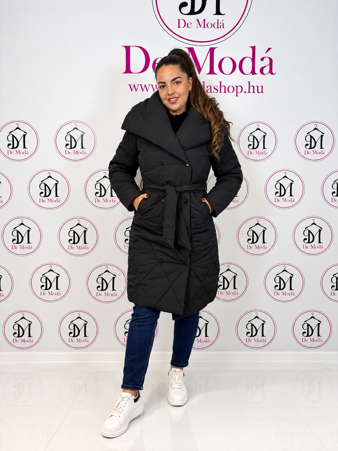 Női modell egy hosszú, fekete téli kabátban, háttérben a De Moda logóival.
