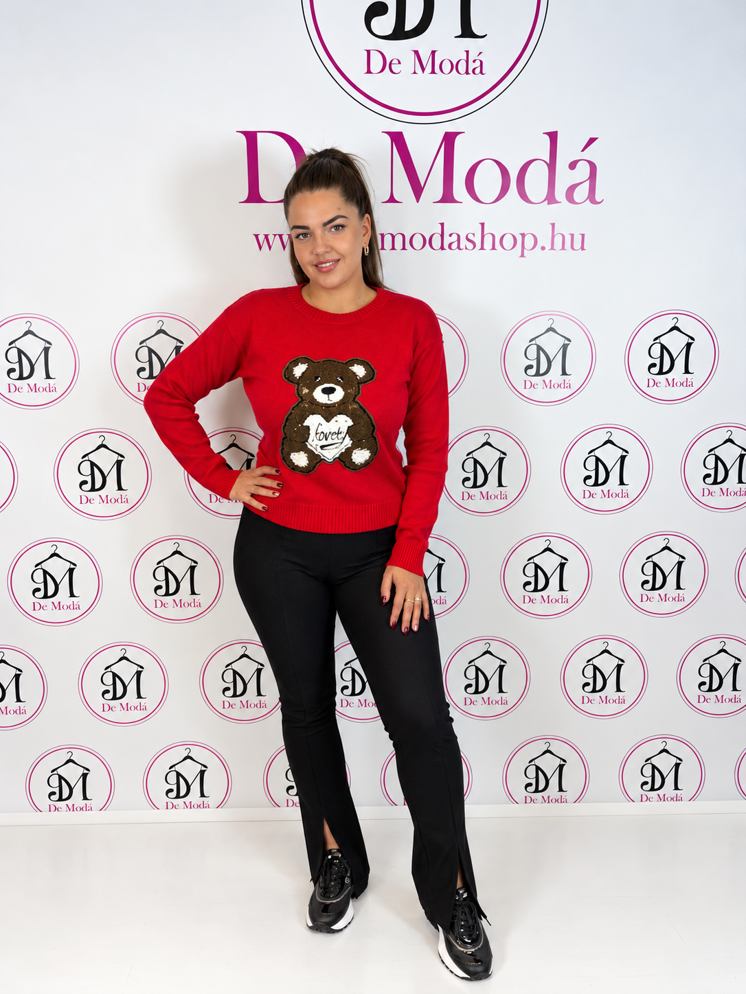Női modell piros macis pulóvert visel, háttérben De Moda logóval.
