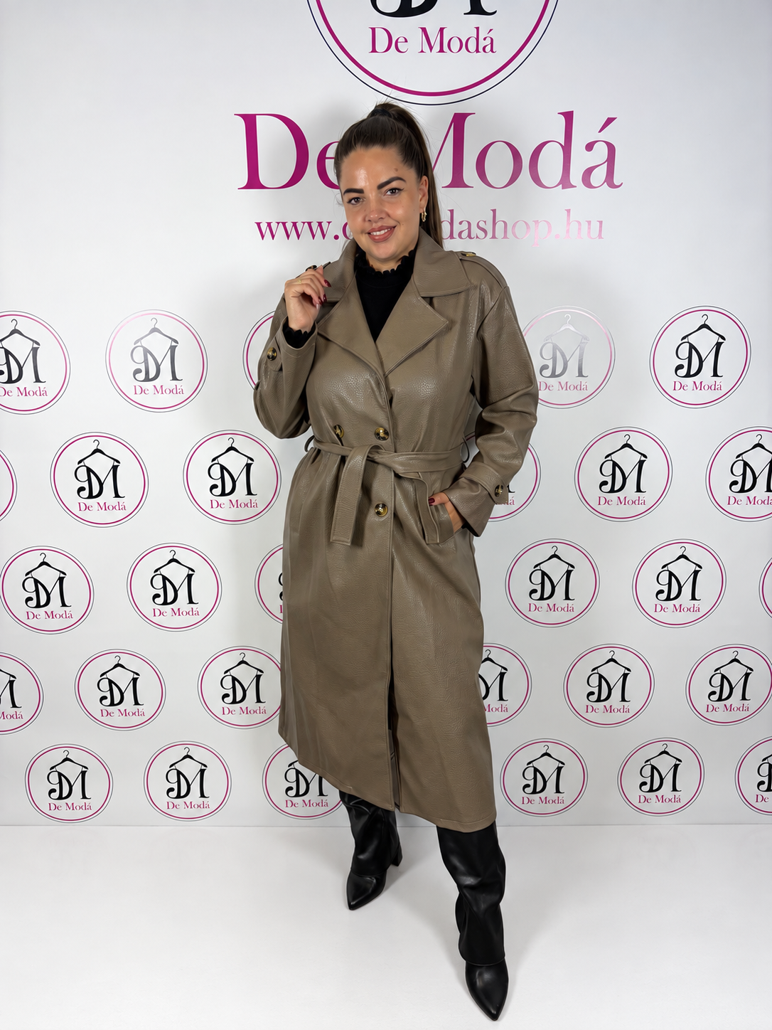 Nő modell hosszú műbőr kabátban, háttérben a De Moda logóval.